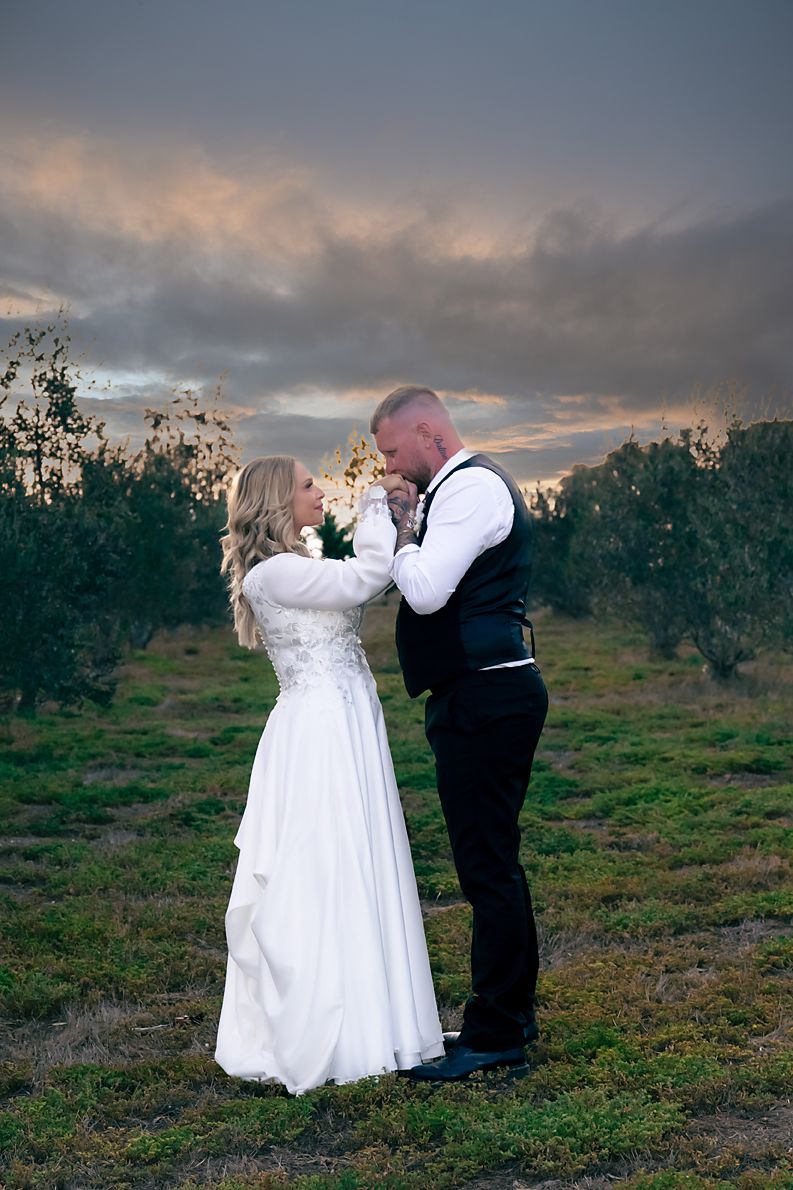A couple in wedding attire stands together in a field at sunset with dramatic cloudy skies in the background.