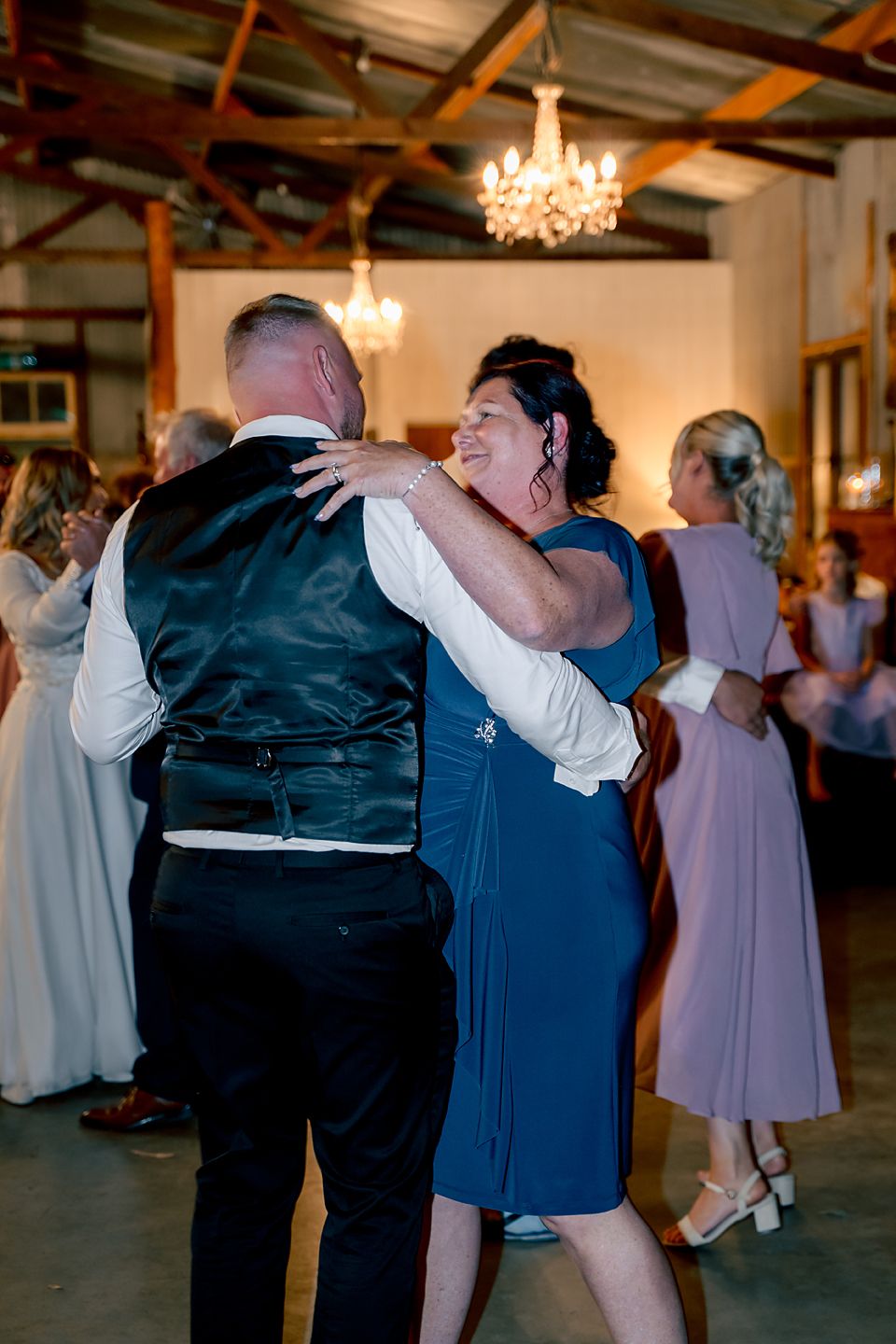 People dancing together at a rustic indoor venue with wooden beams and chandeliers overhead.