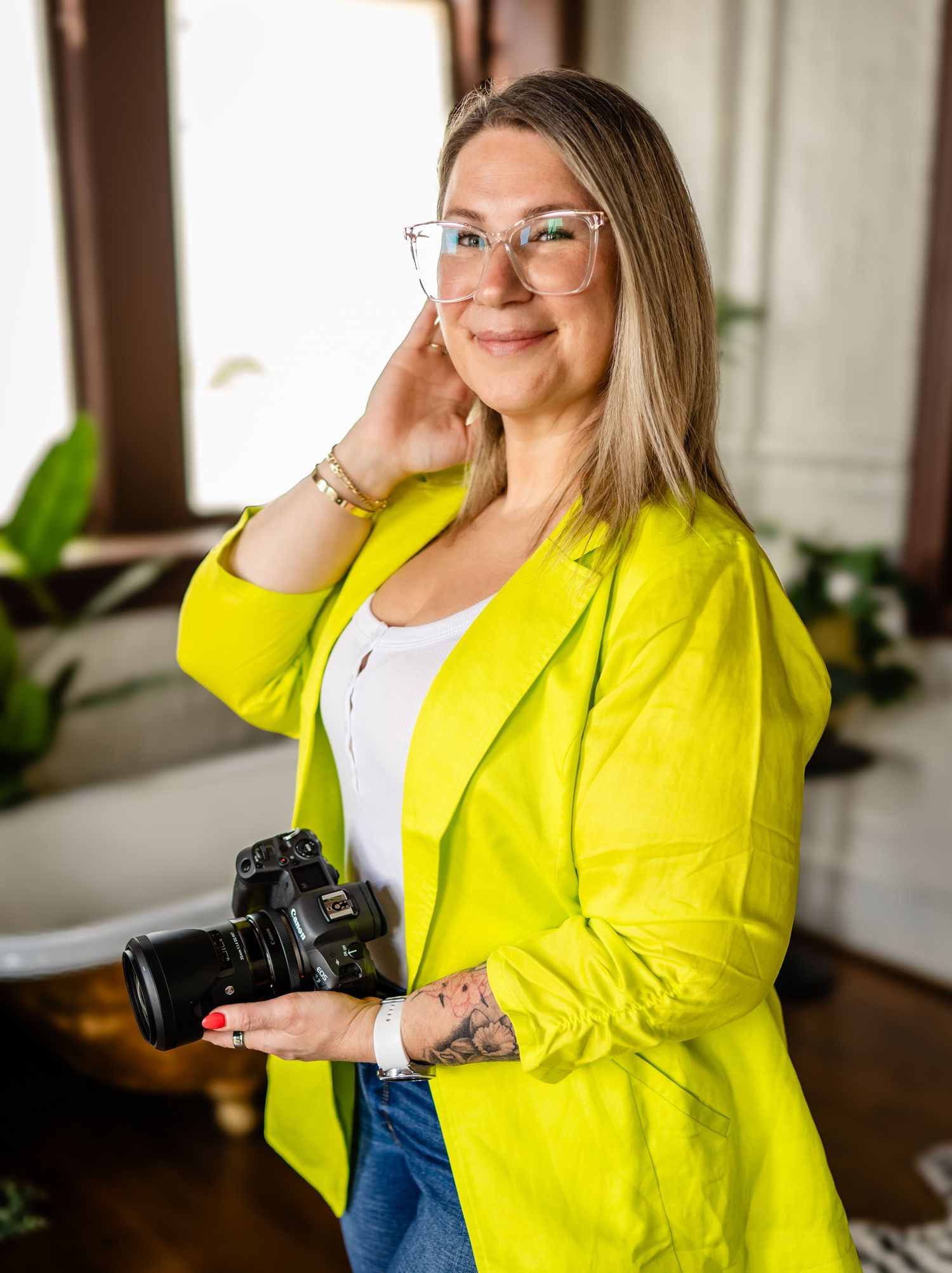 Professional photographer wearing a bright yellow blazer and glasses holds a DSLR camera in a sunlit indoor setting.