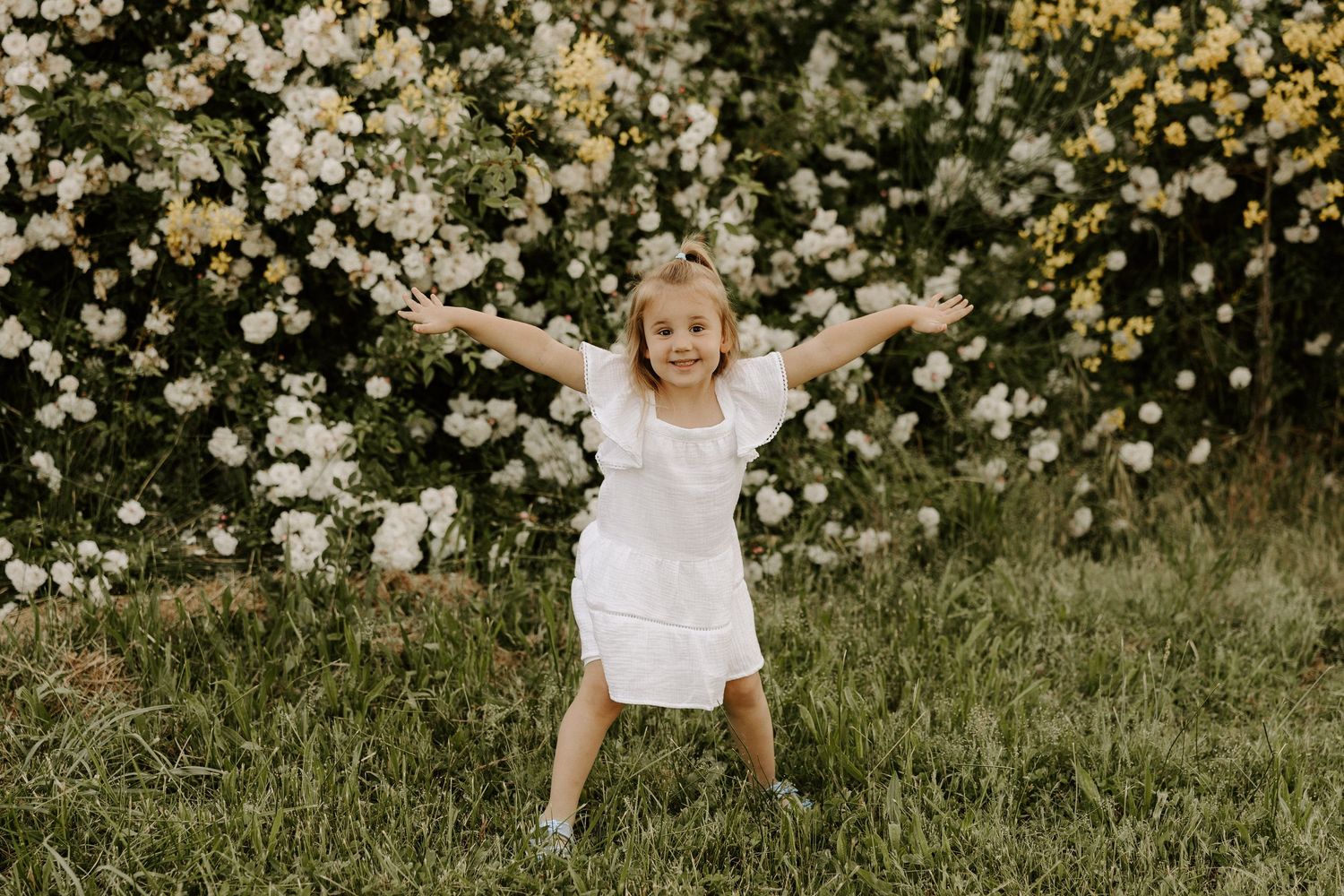 A small child in a white dress jumps joyfully in front of blooming white and yellow flowers.