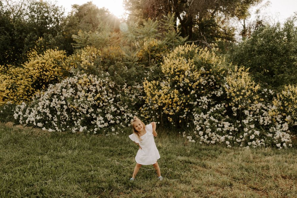 A figure in a white dress twirls in a garden with blooming white and yellow flowers on a sunny afternoon.