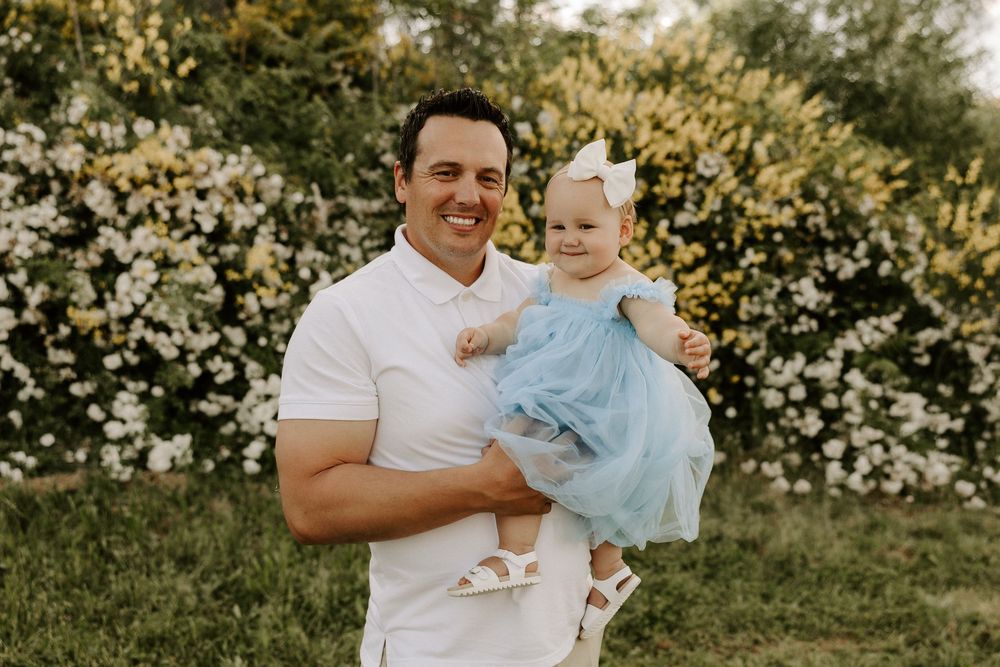 A parent holds a small baby wearing a blue dress while standing in front of blooming white flowers on a sunny day.