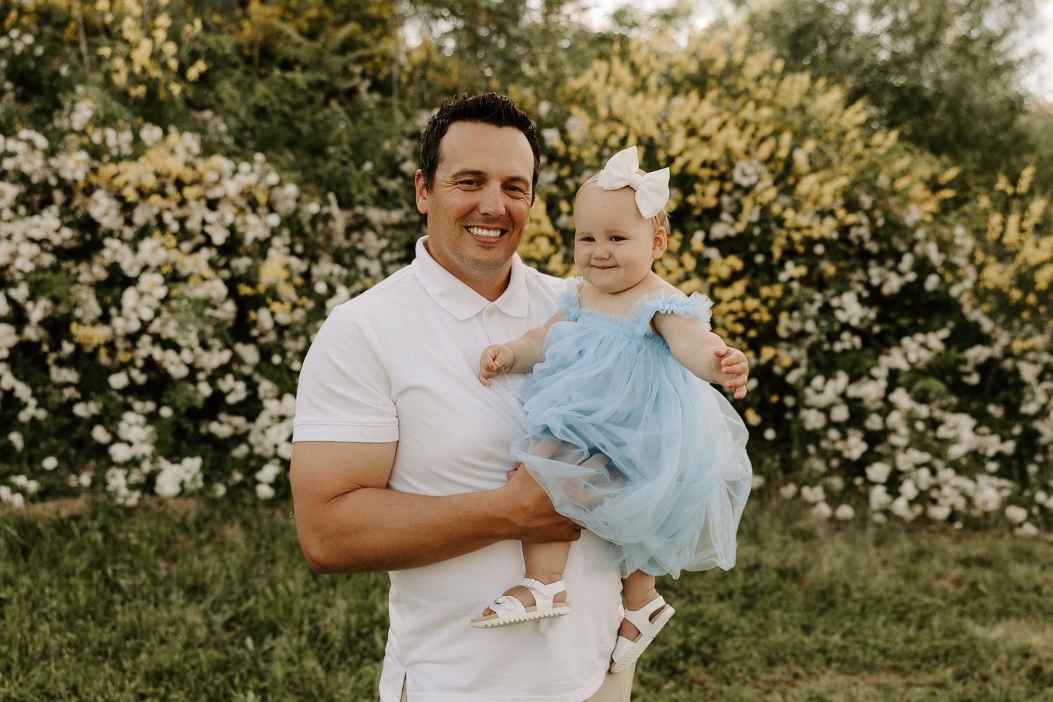 A parent holds a small baby wearing a blue dress while standing in front of blooming white flowers on a sunny day.