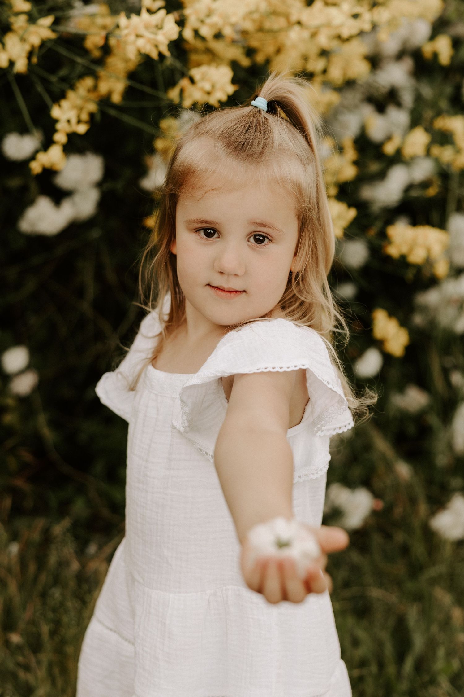 A child in a white dress reaches out while standing in front of blooming yellow and white flowers.