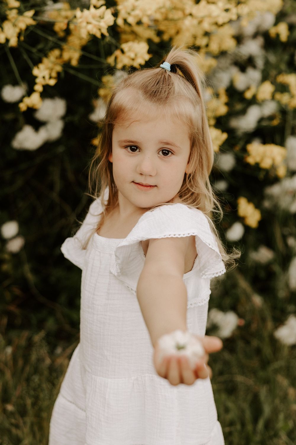 A child in a white dress reaches out while standing in front of blooming yellow and white flowers.