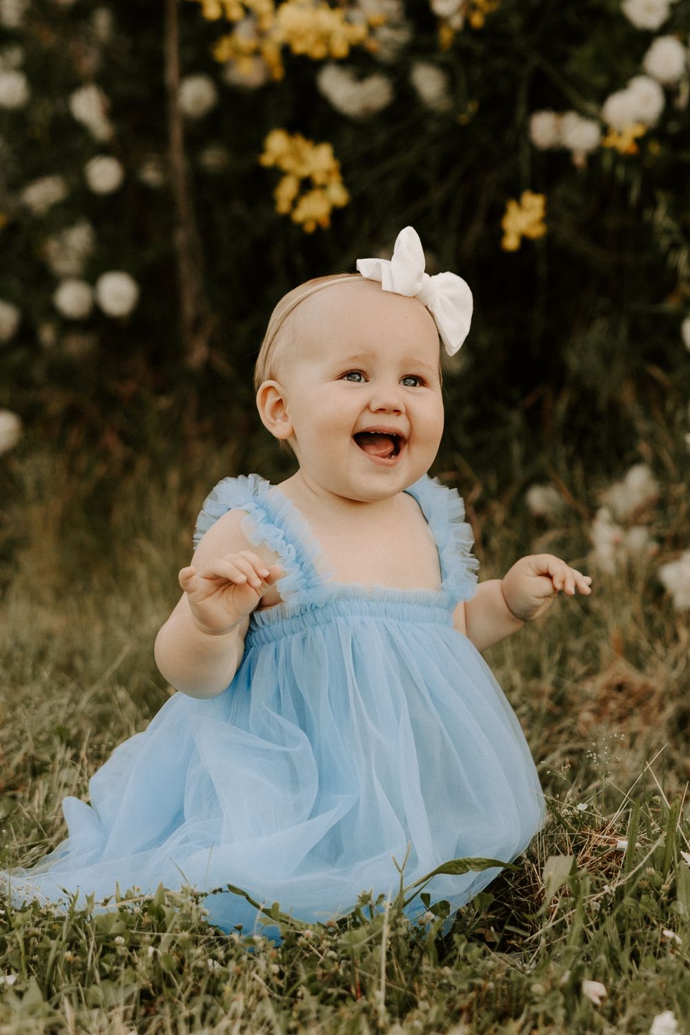 A baby in a blue dress sits on grass surrounded by yellow and white flowers.