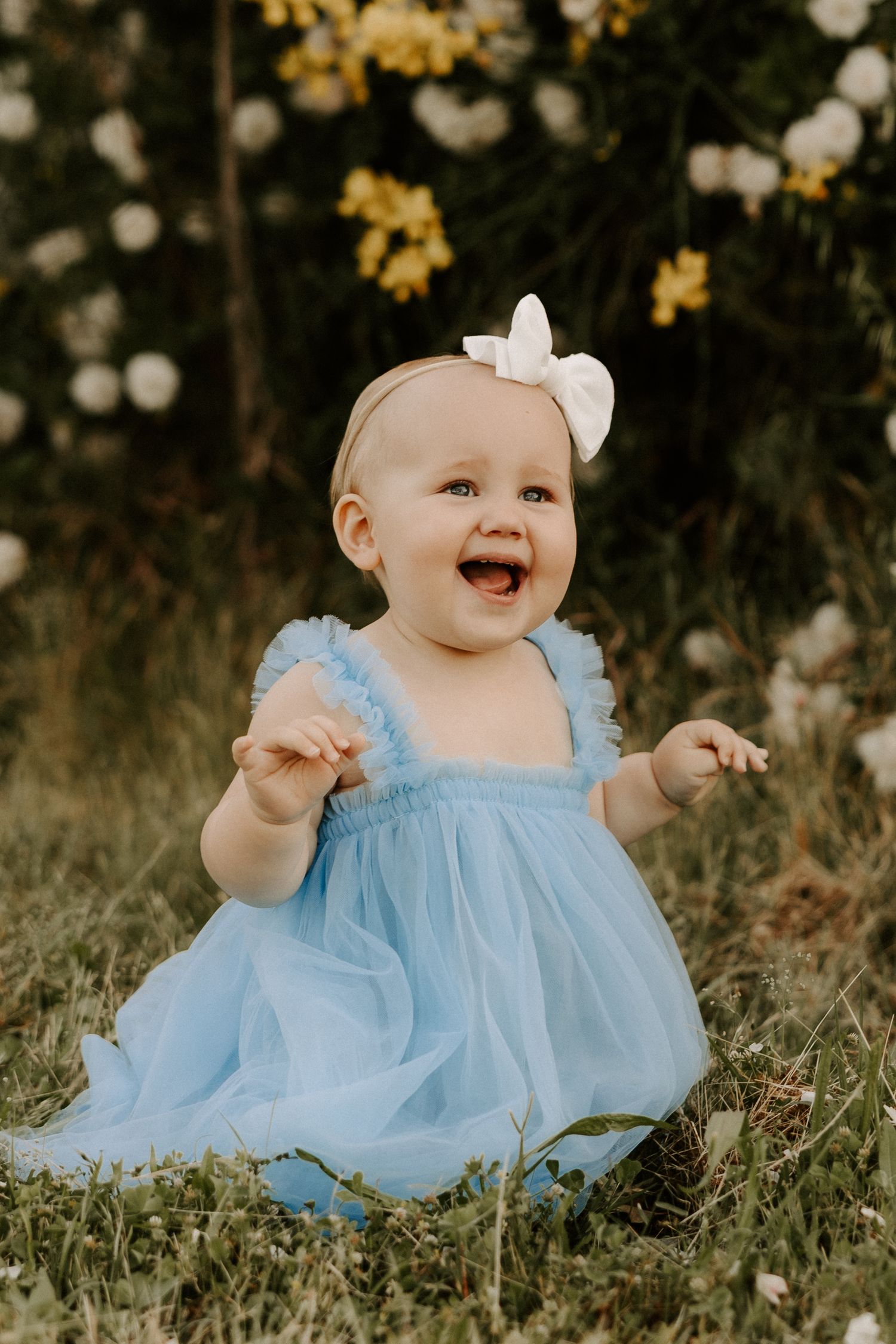 A baby in a blue dress sits on grass surrounded by yellow and white flowers.