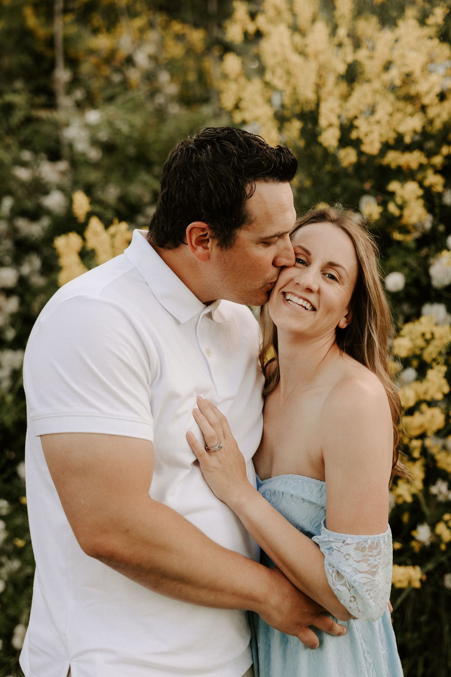 A romantic couple embraces outdoors during a photo session with yellow flowers blooming in the background.