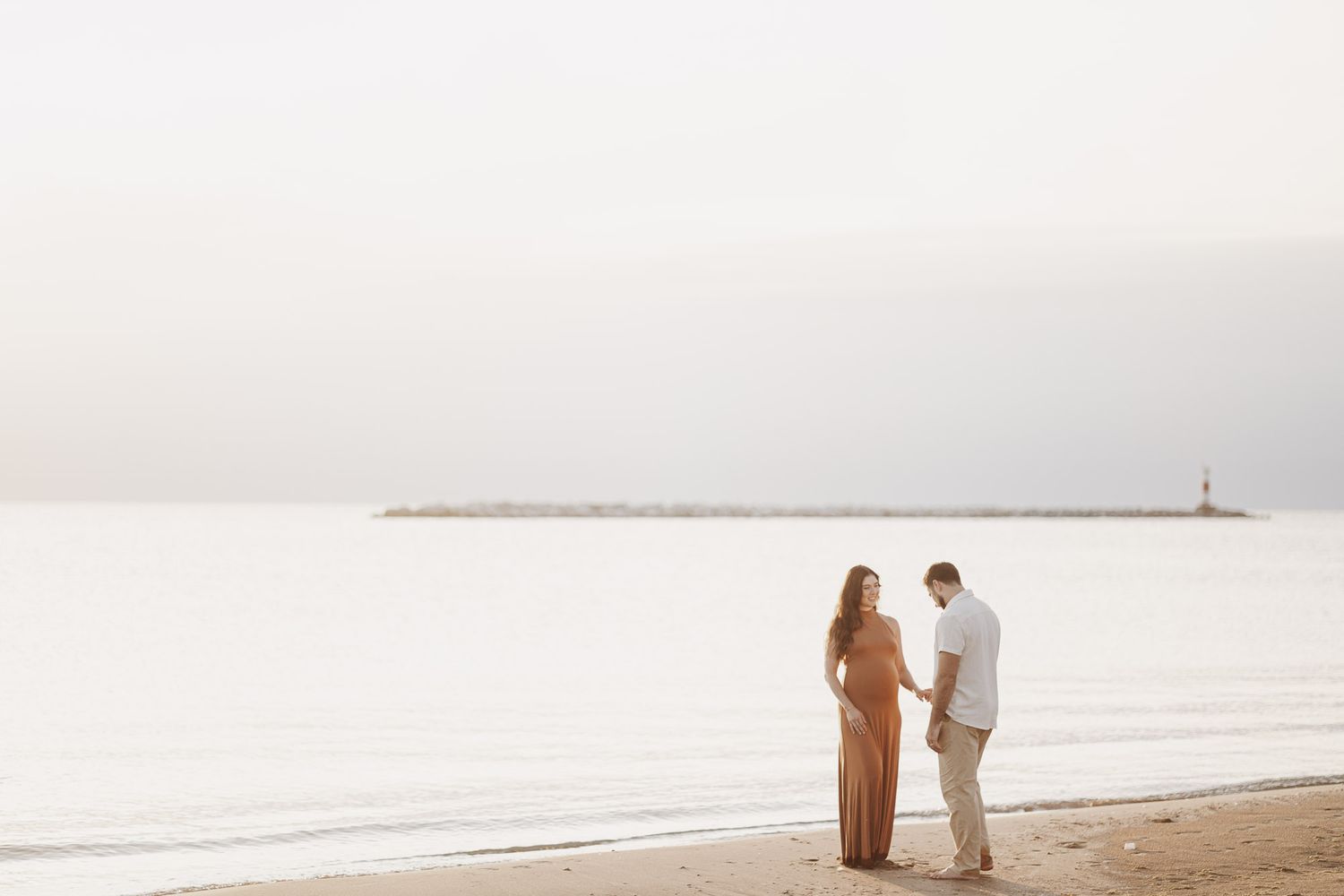 A couple stands together on a serene beach during sunset with a misty horizon in the background at maternity session