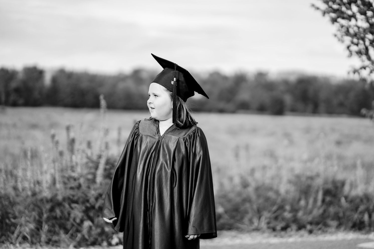 Graduate in black cap and gown stands in a field during a graduation photoshoot session.