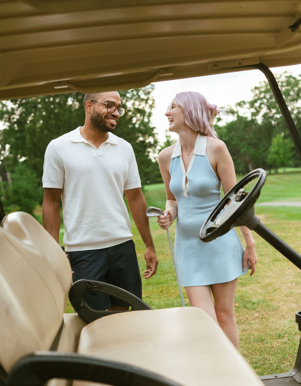 A couple enjoys a casual conversation while leaning against their car in a sunny outdoor setting.