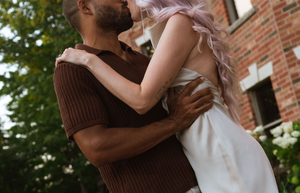 A couple in a brown sweater and white dress embrace lovingly against a brick wall and garden background.