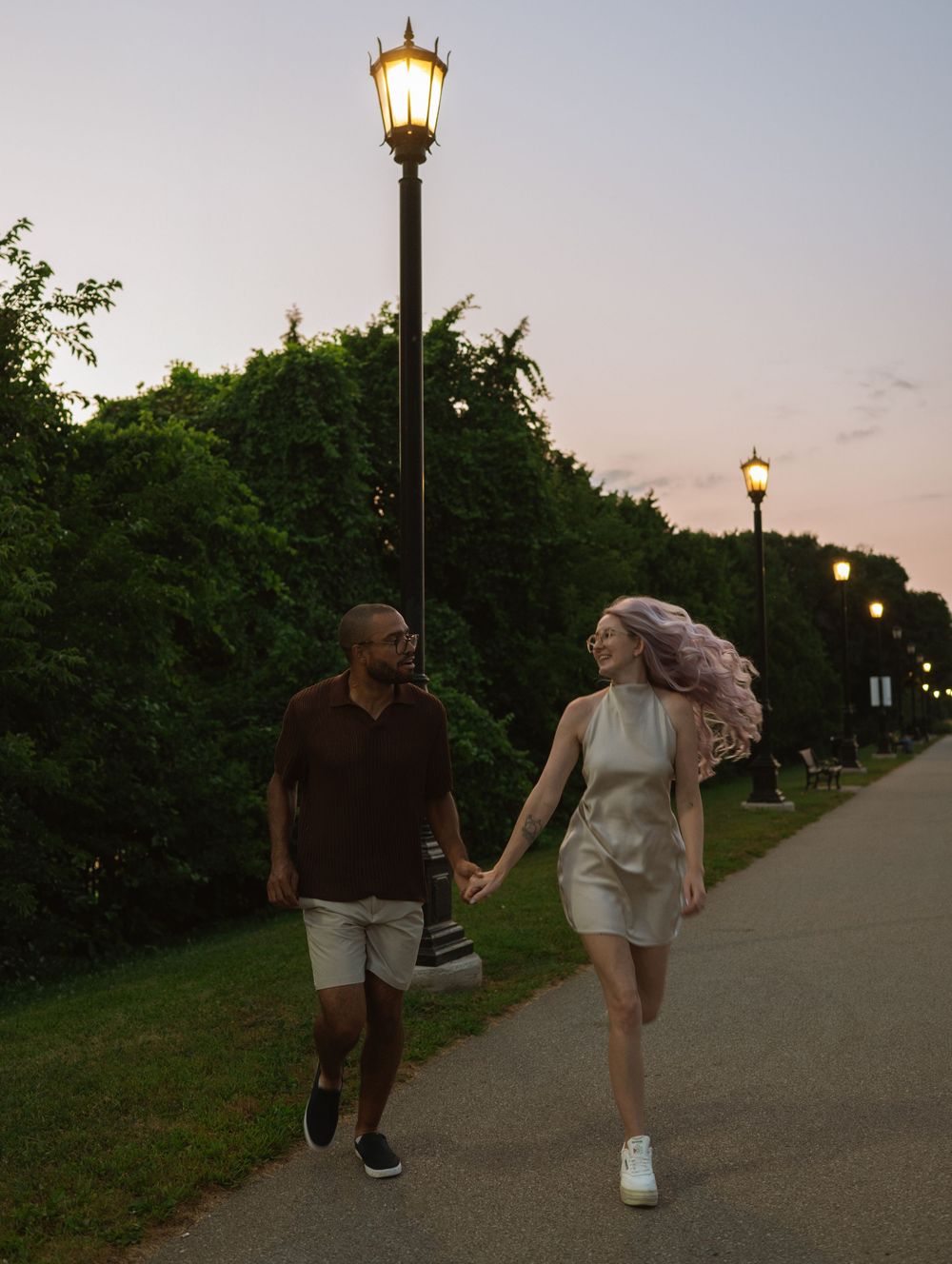 Two people walking together at sunset on a paved pathway with vintage-style lamp posts and green foliage.