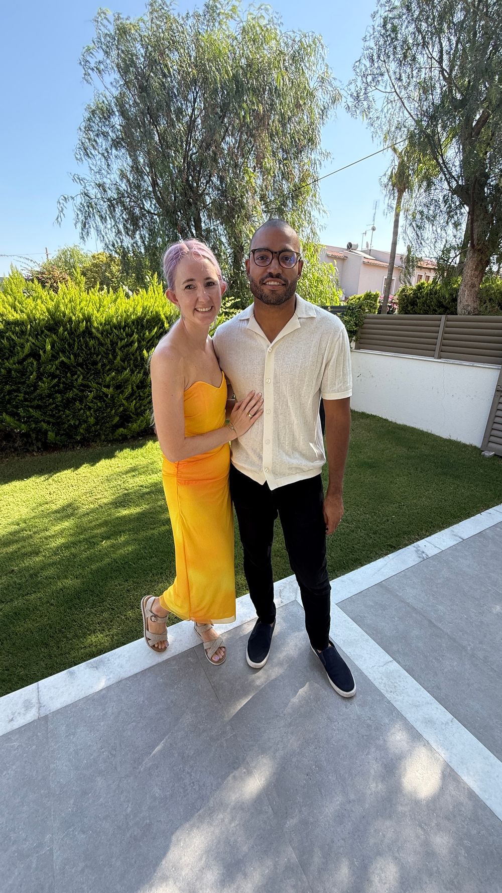 A couple poses together outdoors, with one person wearing a bright yellow dress and the other in a white shirt.