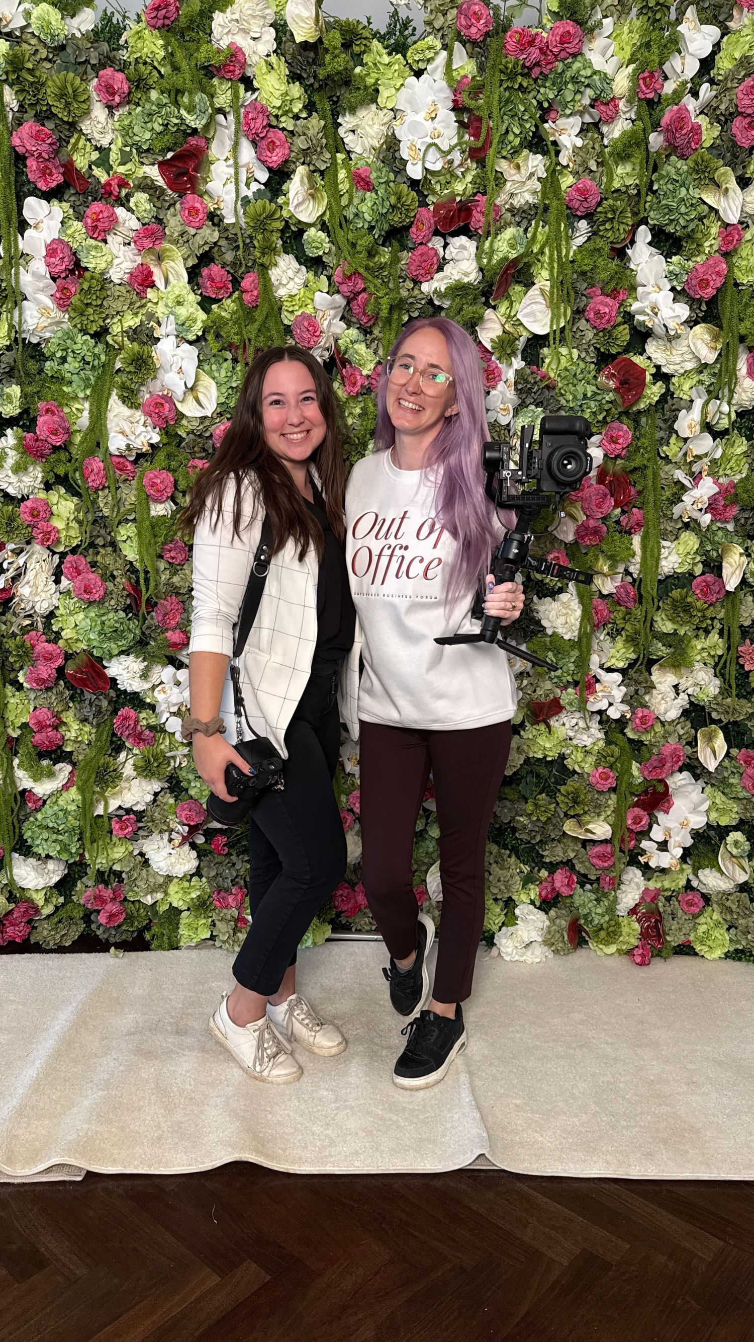 Two friends pose in matching outfits against a beautiful floral wall backdrop with pink and white roses.