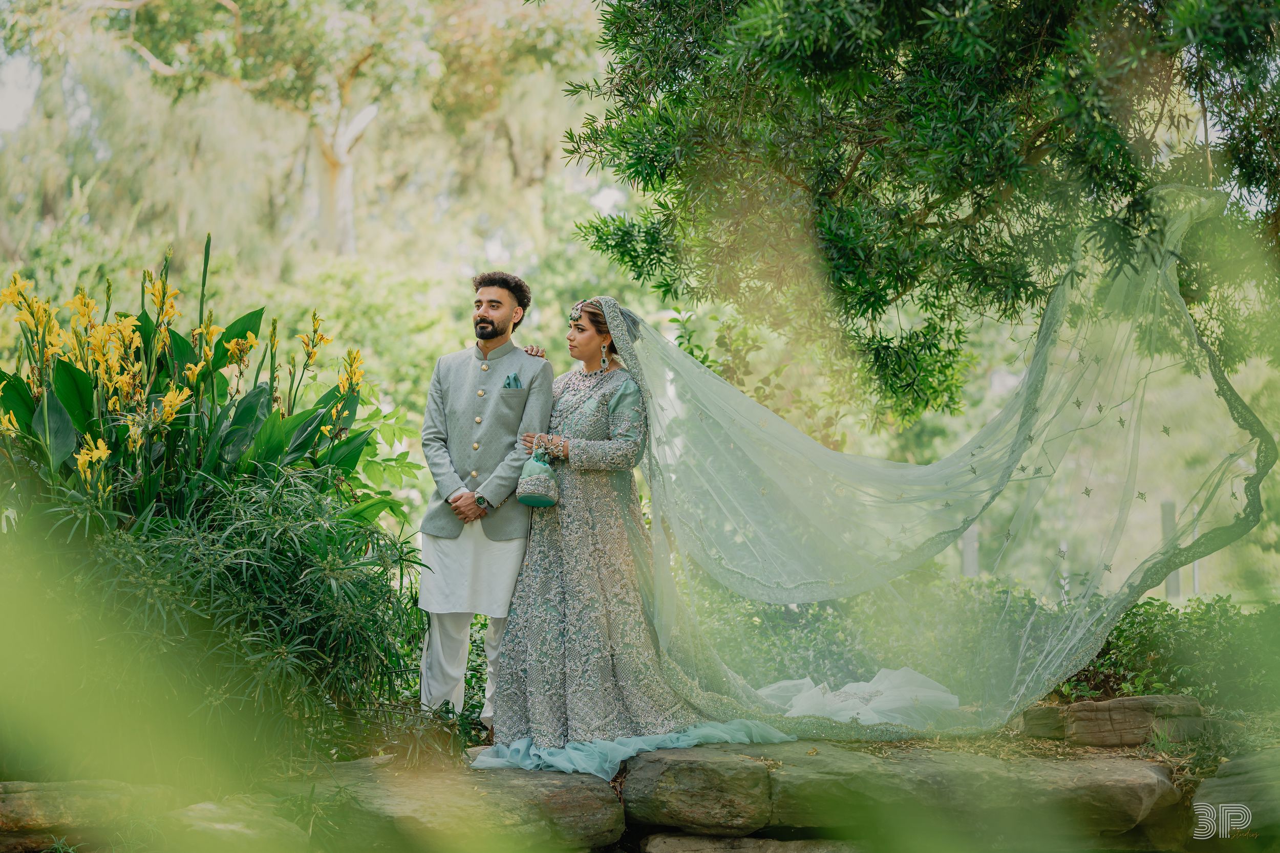 A couple in wedding attire poses together in a lush garden setting with flowing veil and natural lighting.