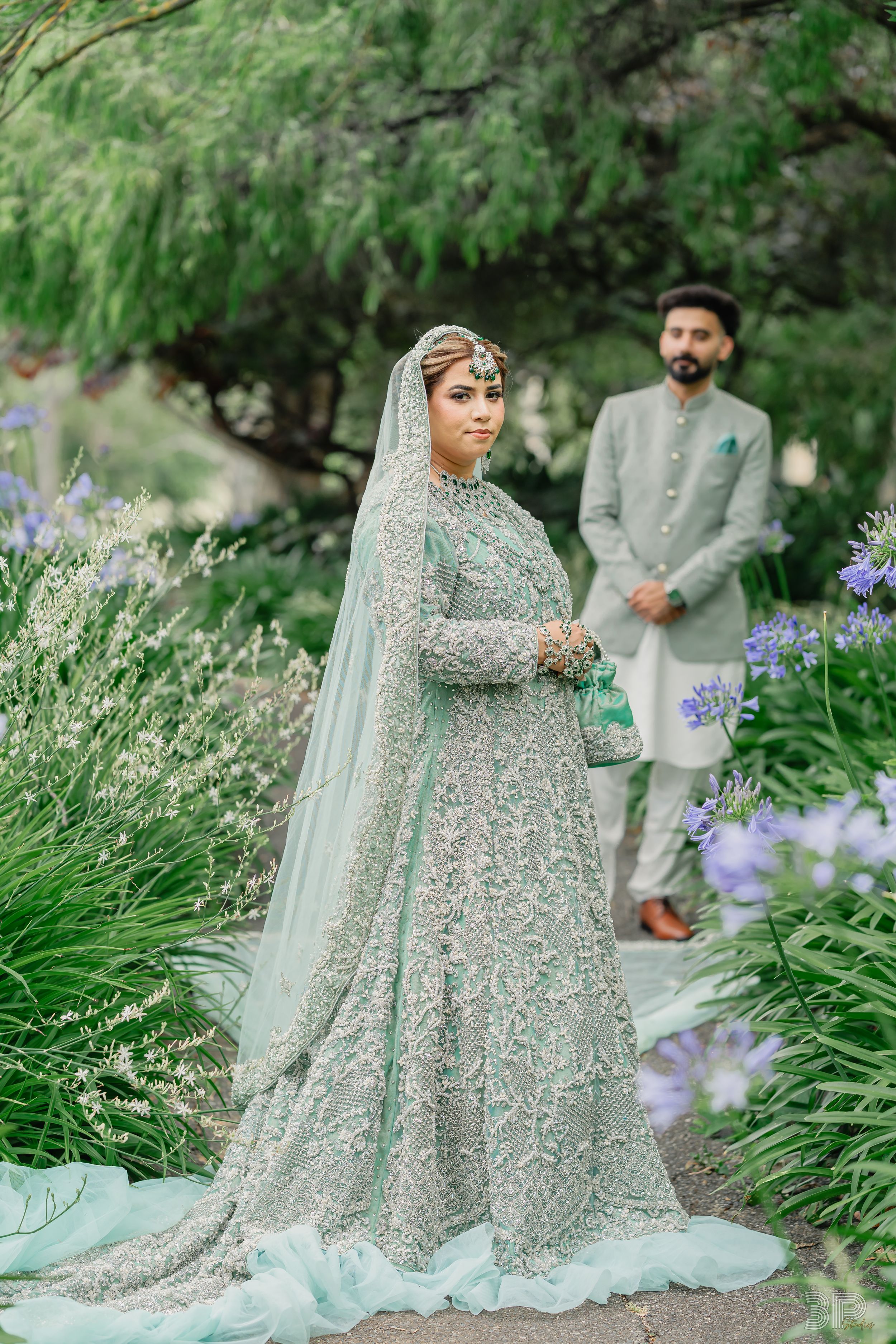A couple in traditional South Asian wedding attire poses in a garden with blooming purple iris flowers.