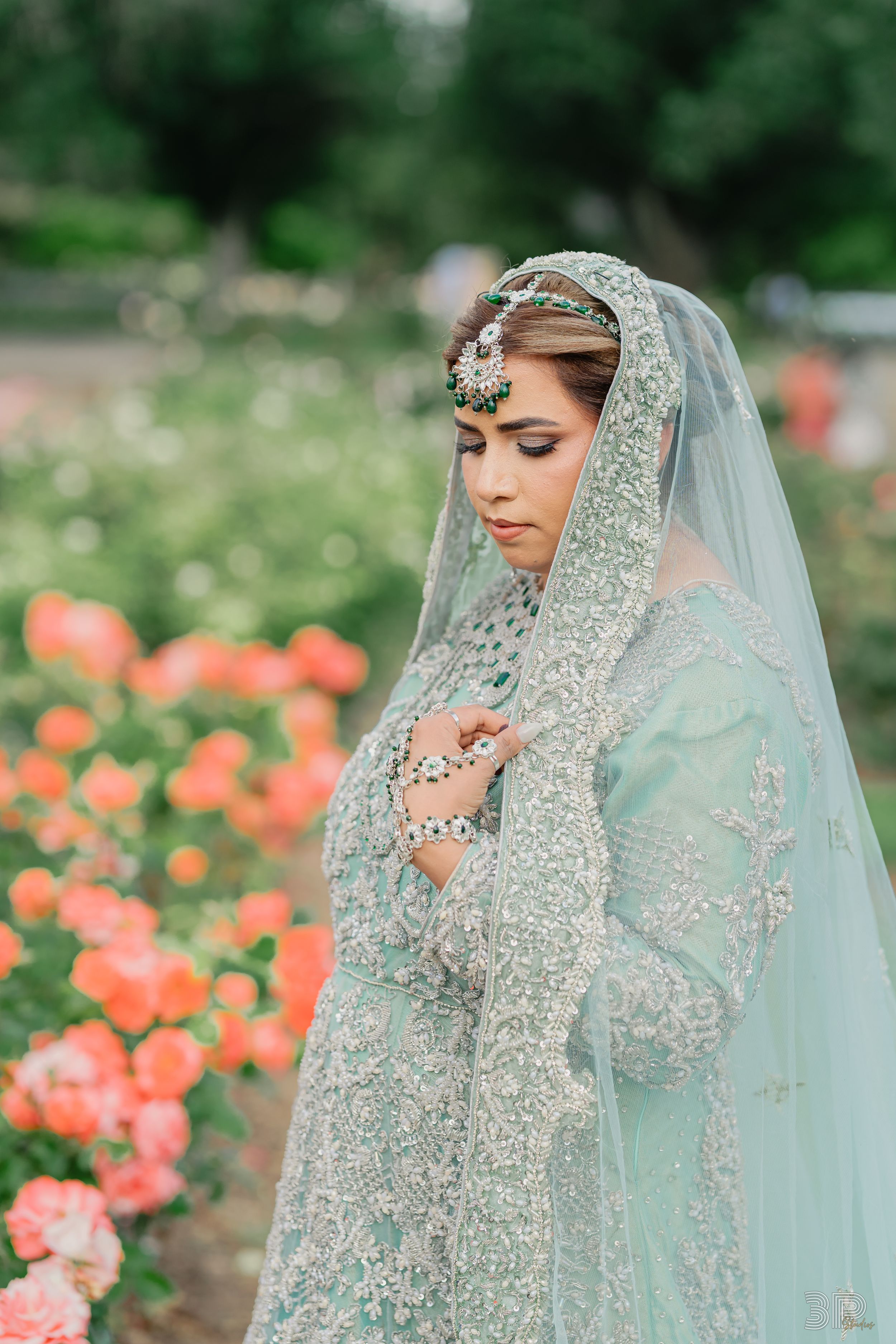 Bride wearing an elegant mint green and silver embellished wedding dress with matching veil in a garden of coral roses.