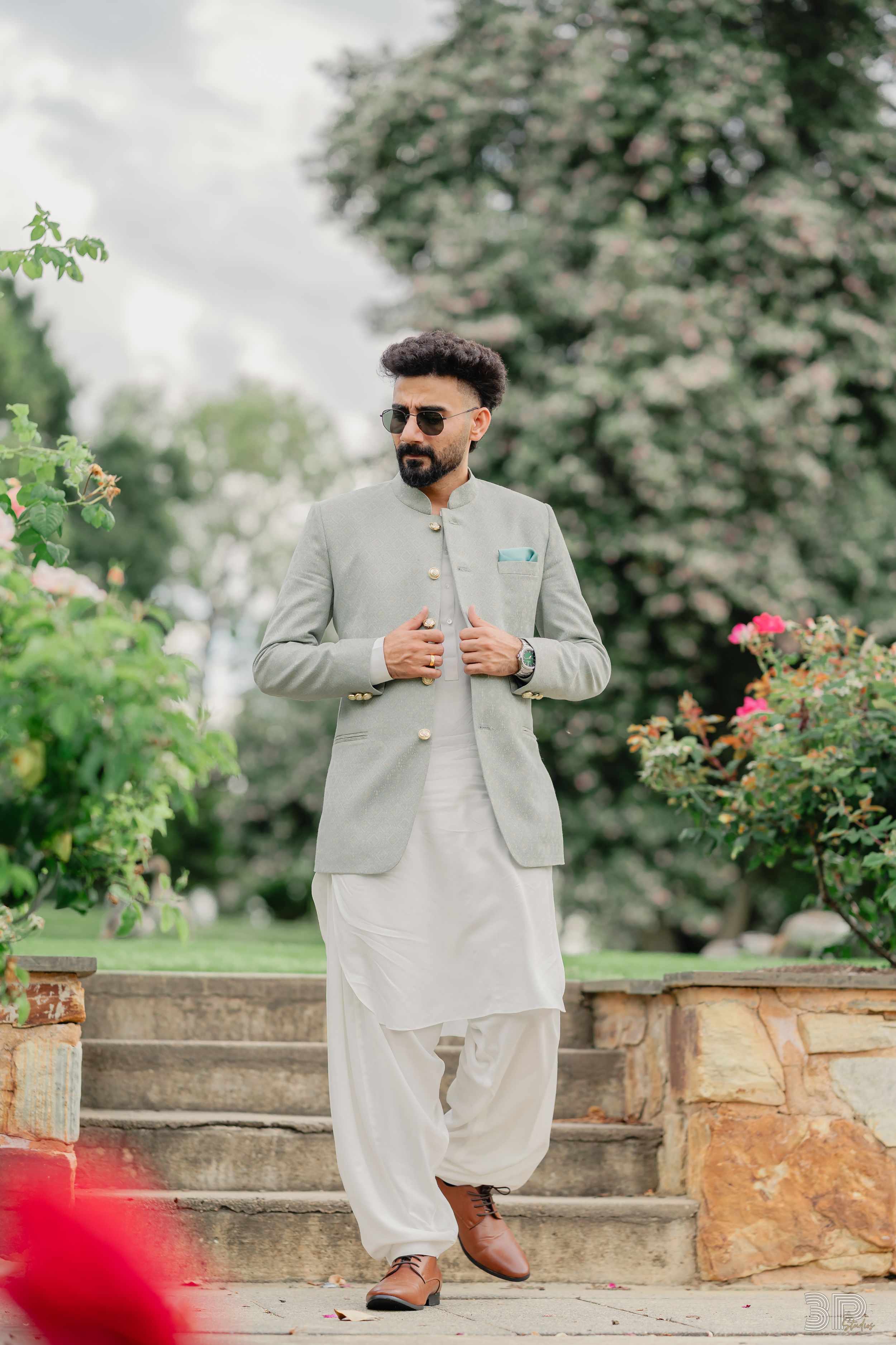 A person in traditional white shalwar kameez and grey blazer poses in a garden setting with stone walls and flowers.