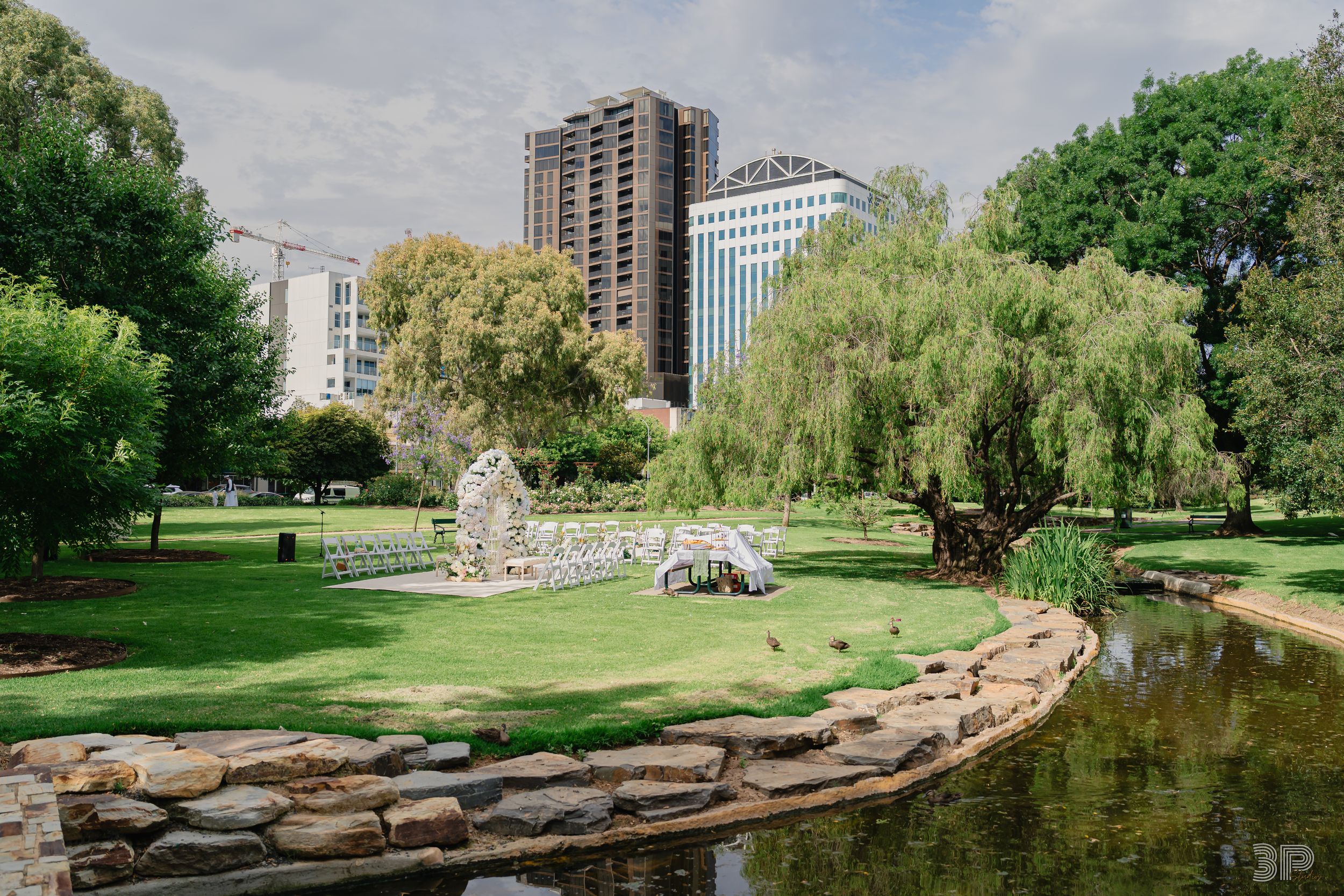 A scenic panoramic view of a lush public park with stone walls, ponds, and tall buildings in the background.