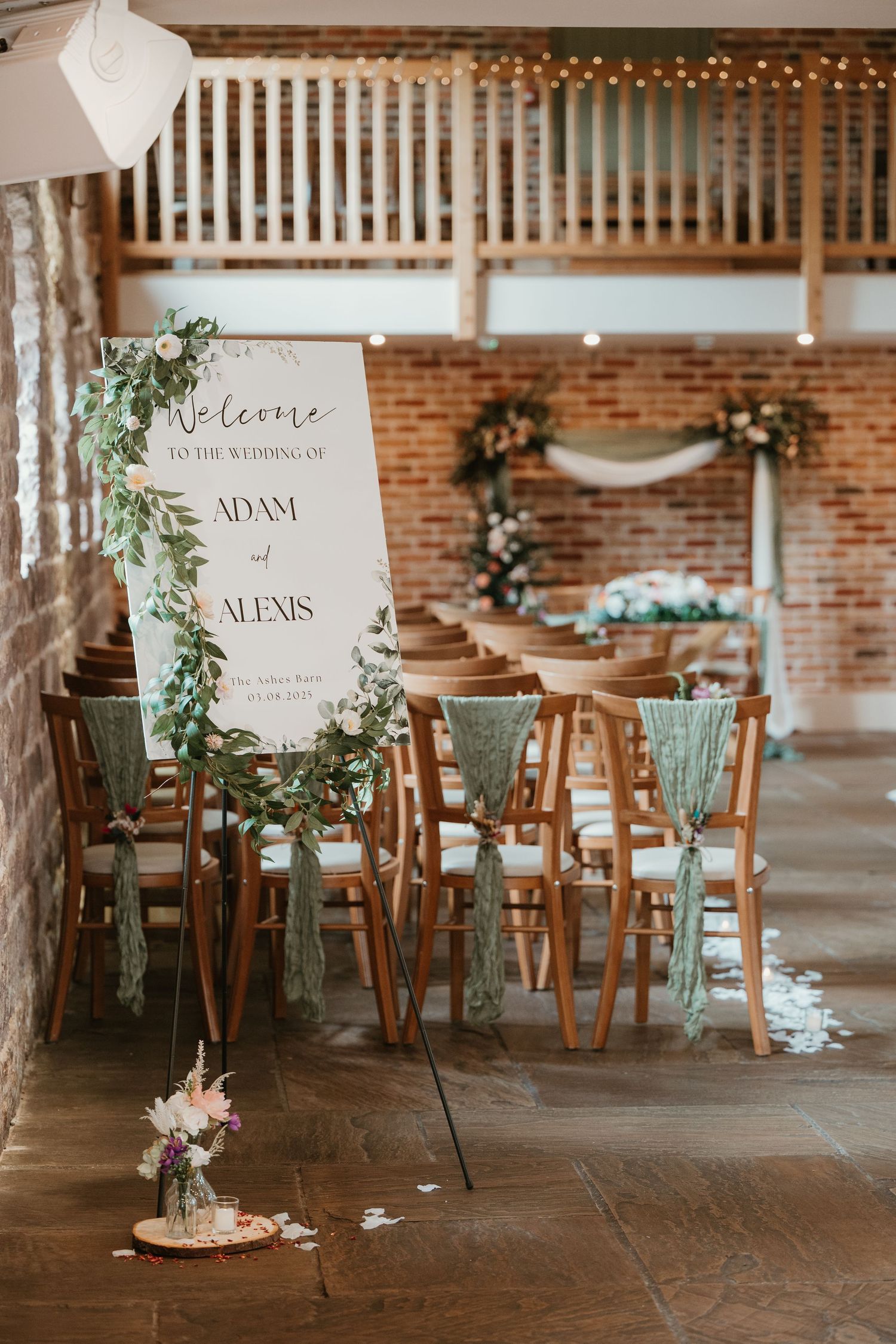 Rustic wedding reception setup with wooden chairs, greenery decorations and personalized name signs in a brick-walled venue.