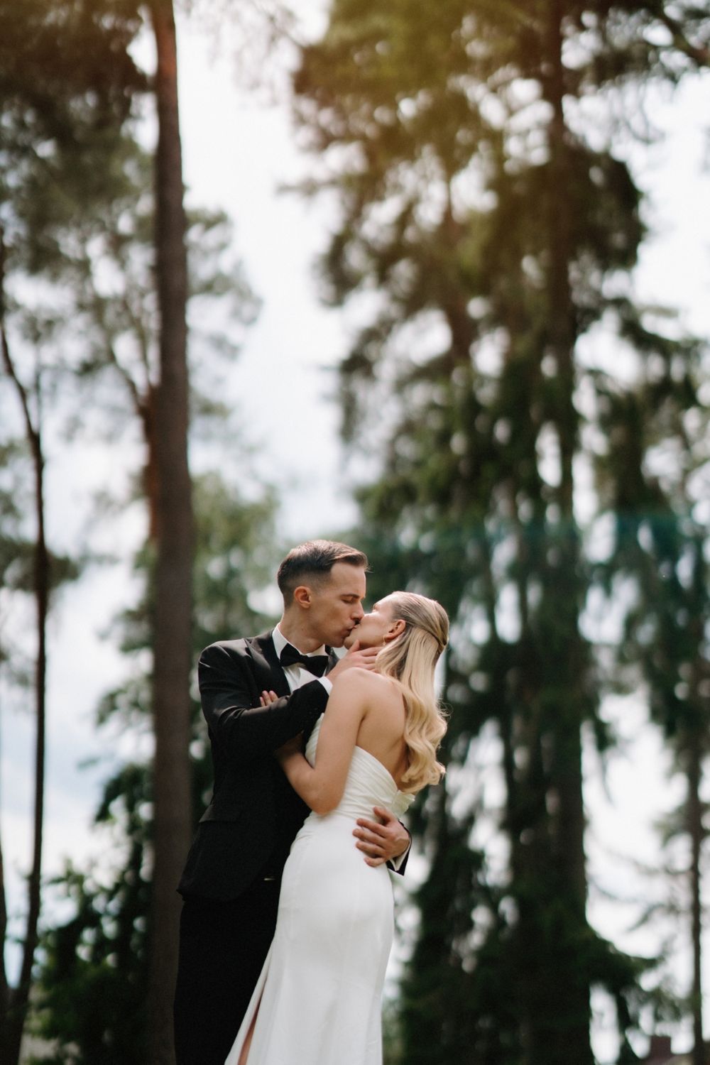 Romantic wedding portrait of couple embracing under tall evergreen trees.