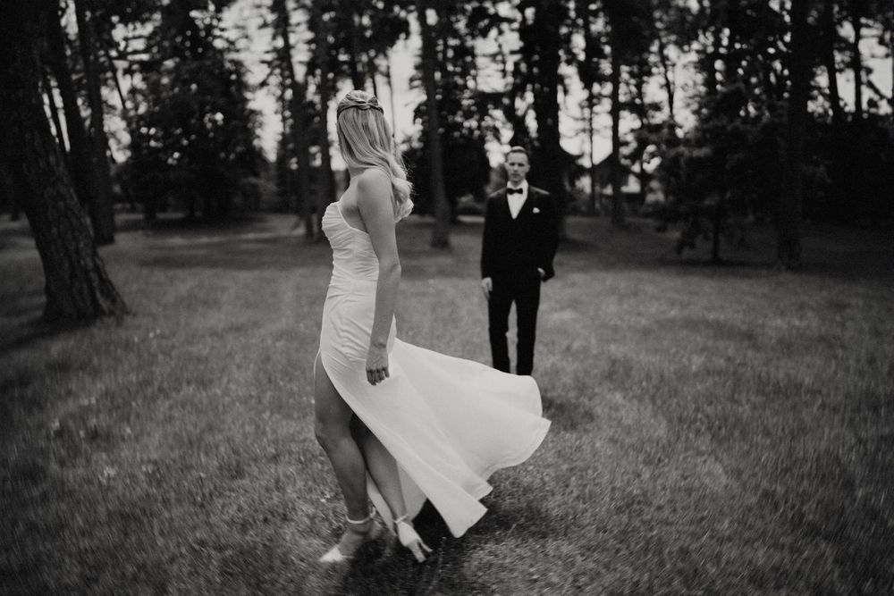 Black and white photo of bride's dress flowing in breeze as groom watches from distance.