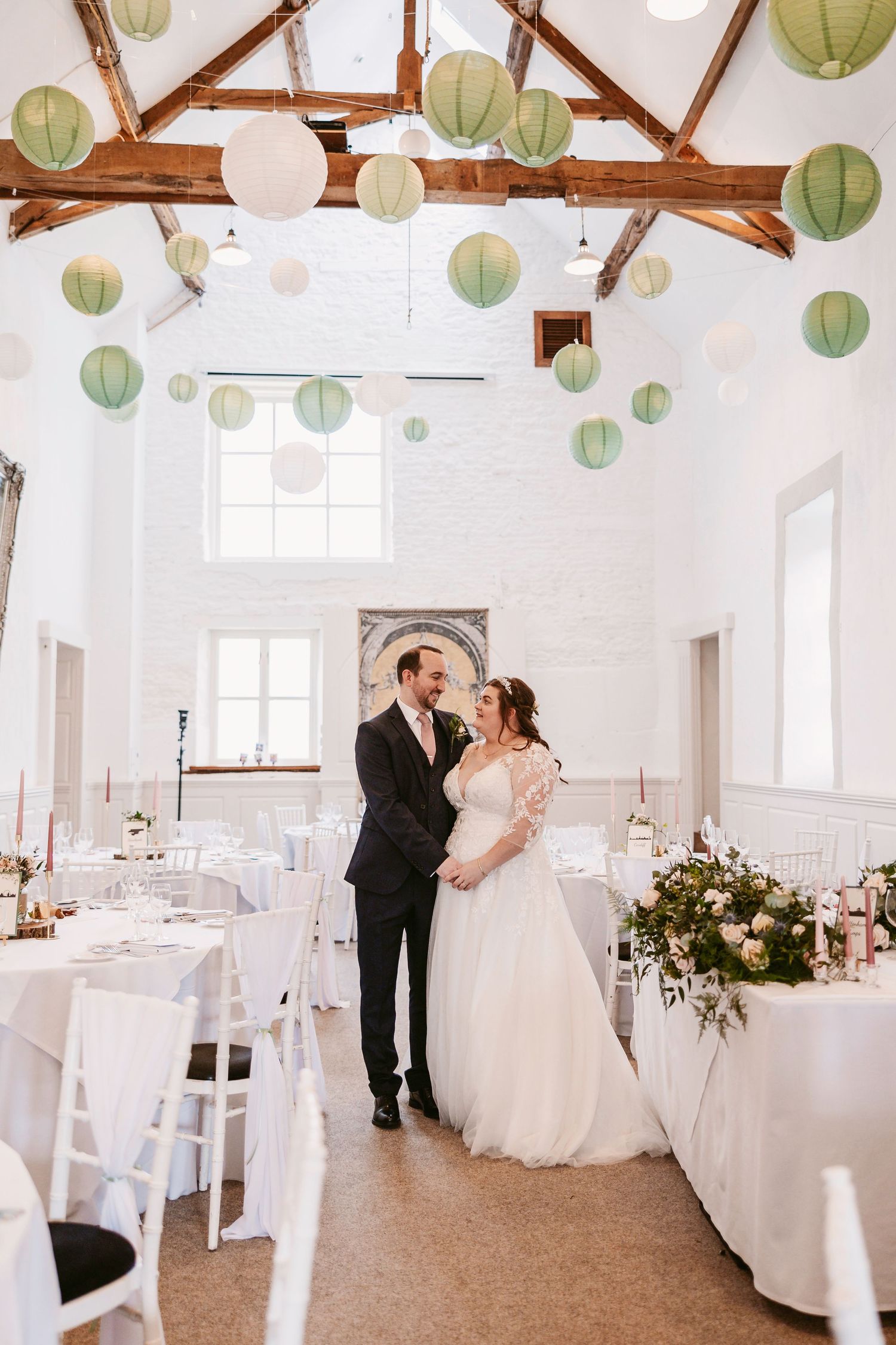 Elegant white wedding reception hall with exposed wooden beams and hanging paper lantern decorations.