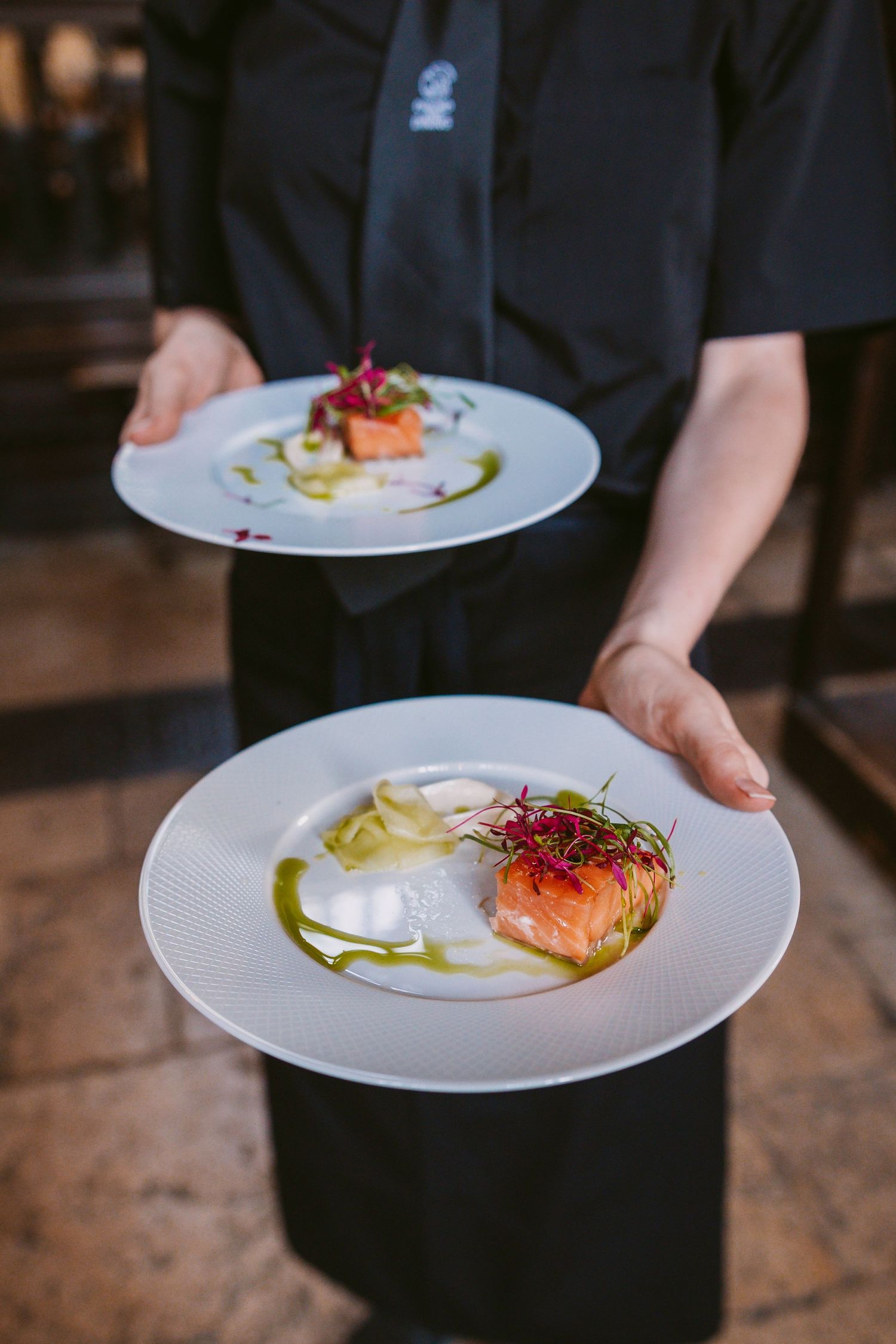 Servers holding white plates with beautifully plated salmon dishes garnished with microgreens and colorful sauces.