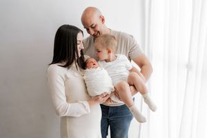 Parents in white and gray clothing share tender moments with their newborn baby against a bright white backdrop.