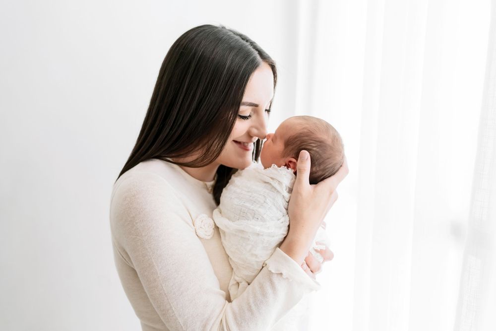 A parent in a white sweater lovingly holds and kisses a newborn baby against a bright white background.