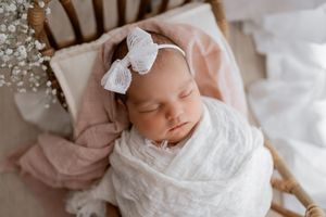 A baby wearing a white lace dress and bow headband rests peacefully on soft white bedding.