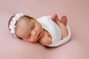 Peaceful newborn baby sleeping on pink backdrop wearing white wrap and delicate floral headband.