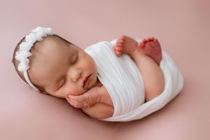 Peaceful newborn sleeps on pink backdrop while wrapped in white swaddle cloth.