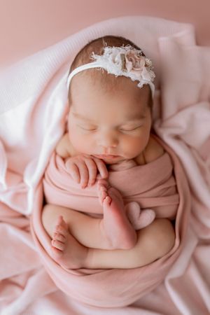 Sleeping newborn baby wrapped in soft pink fabric with delicate white lace headband in professional studio portraits.