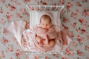 Newborn baby wrapped in soft pink fabric lying in white crib with floral background.