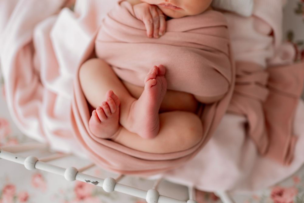 Close up of tiny baby feet and hands wrapped in delicate pink swaddle on white bed.