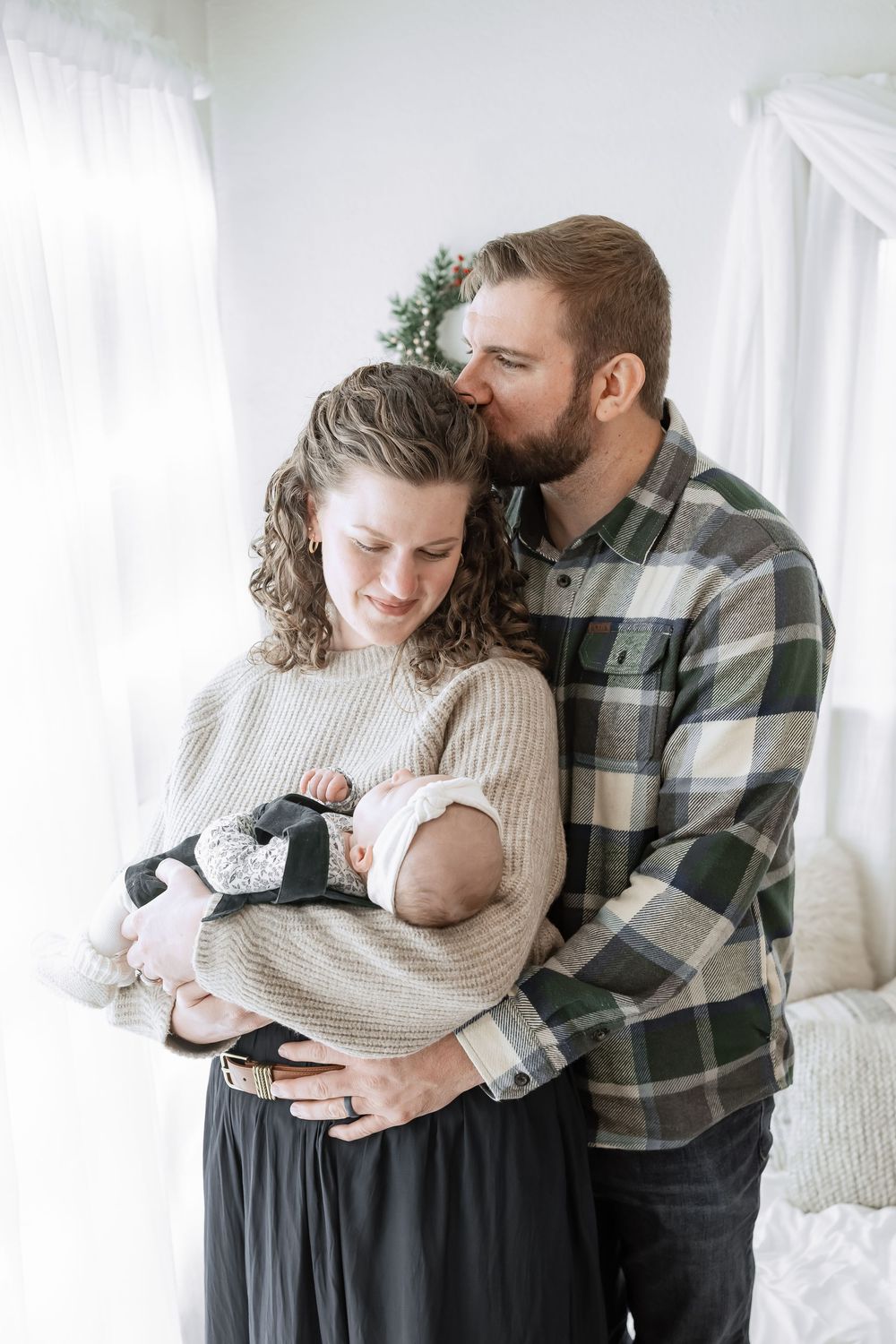 A couple embraces while holding their newborn baby in a bright white room with natural light.