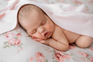 Newborn baby rests on floral patterned fabric with soft pink background and delicate lighting.