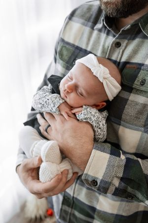 A sleeping newborn baby wearing a white lace dress and bow headband is cradled in plaid-shirted arms.