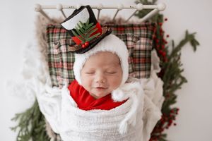 A sleeping baby wearing a white knit hat and swaddle is surrounded by festive Christmas decor and plaid fabric.