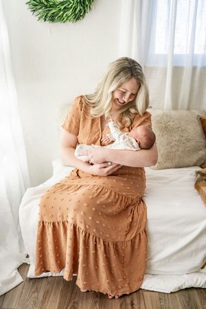 A mother in a peach-colored dress sits on a white couch holding and bonding with her newborn baby in a bright room.