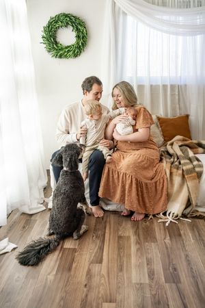 A couple sits on hardwood flooring at home with their large black dog in a bright room with white curtains.