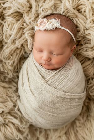 A sleeping newborn wrapped in soft cream fabric lies on a textured beige carpet in a peaceful newborn photography setup.