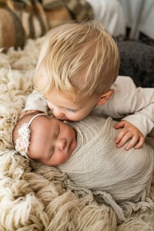 A tender moment captured as a young child gently kisses a sleeping newborn swaddled in soft gray fabric.