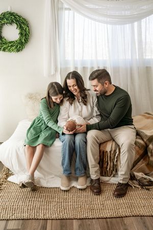 A family sits together on a couch with a newborn baby beneath white curtains and a green wreath on the wall.