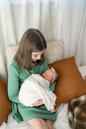 A person in a green sweater holds and gazes lovingly at a sleeping newborn baby wrapped in white blankets.