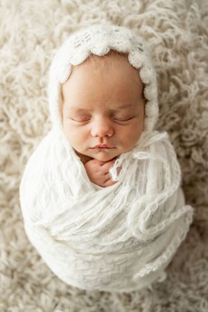 Sleeping newborn wrapped in soft white lace bonnet and swaddle on textured cream background.