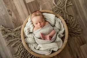 Newborn baby sleeping peacefully in round wooden bowl wrapped in soft gray knitted blanket.