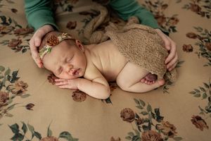 Sleeping newborn on floral patterned fabric wearing tan knit wrap with hands gently cradling.